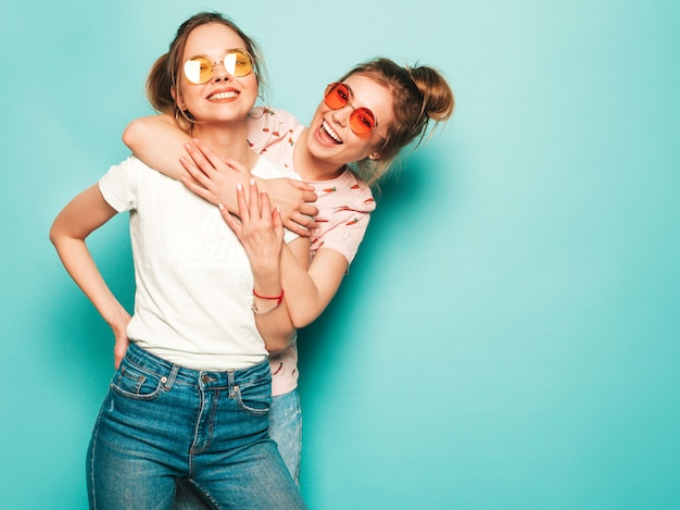 Free photo two young beautiful blond smiling hipster girls in trendy summer hipster jeans clothes. sexy carefree women posing near blue wall. trendy and positive models having fun