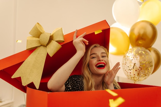 Joyful fairskinned young blonde enthusiastically climbs out of large red box as surprise holiday concept